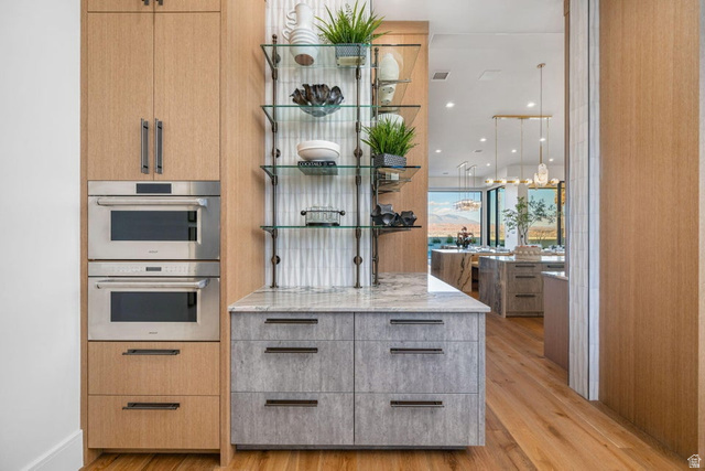 2867 East Granite Way St. George, UT 84790 - Photo 18 of 69 Built-in stainless steel double ovens, custom wood-finish cabinetry, gray textured lower cabinetry with dark hardware, glass shelving with dark metal supports, and a light-toned countertop