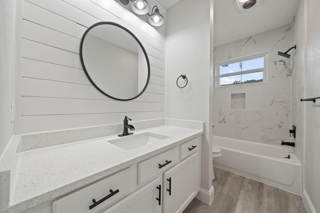 310 Valley Spring Drive Arlington, TX 76018 - Photo 20 of 24 Bathroom featuring a white shiplap accent wall, white vanity with a light-toned countertop, black hardware and fixtures, a built-in tub with marble-look tile surround, and wood-finish flooring