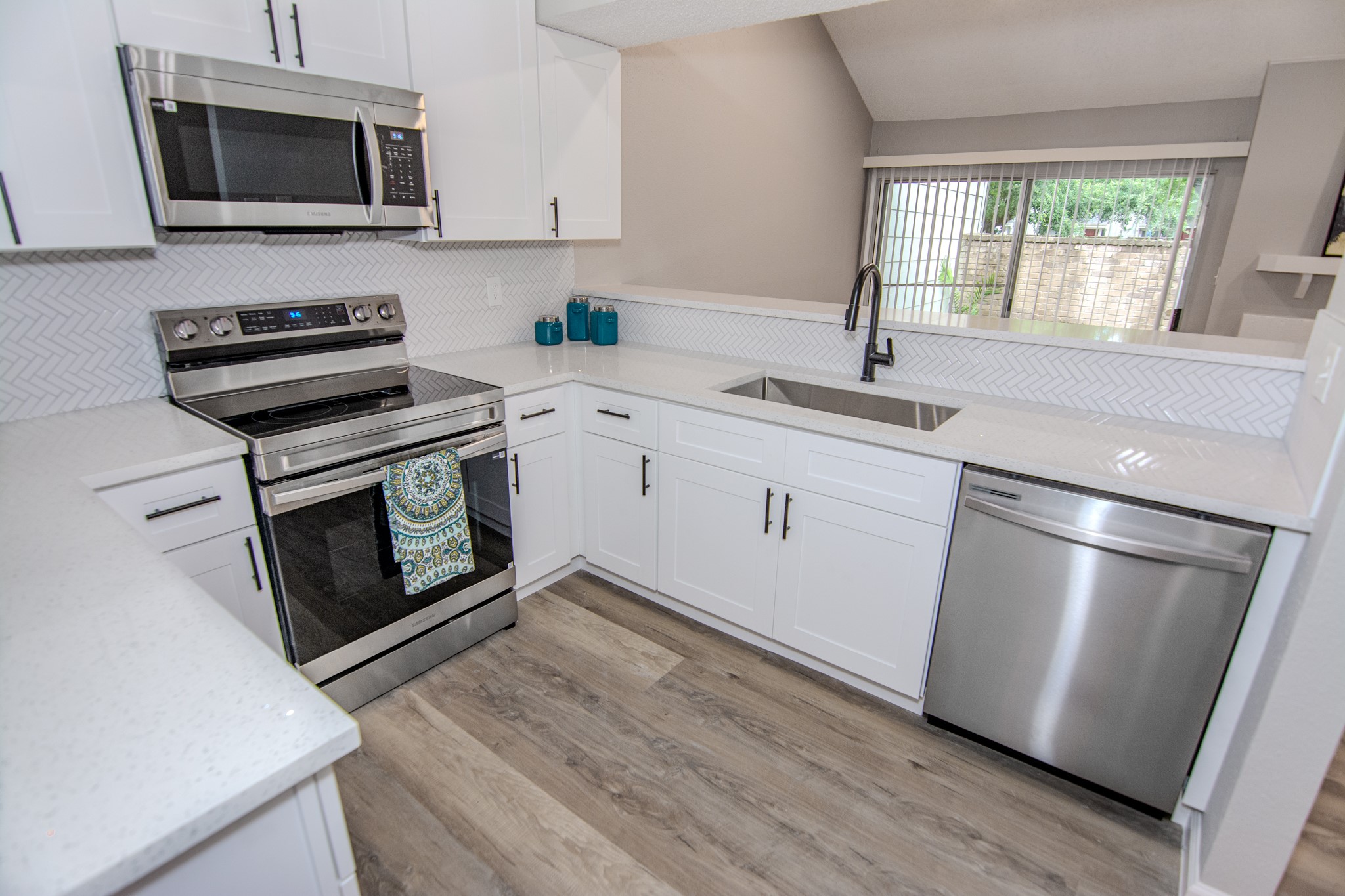 13009 Wirevine Lane Houston, TX 77072 - Photo 2 of 18 Modern kitchen with sleek white cabinets, stainless steel appliances, and a stylish herringbone backsplash. The open layout features a large sink, ample counter space, and a view towards the patio.