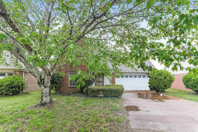 a sign that is sitting in front of a house with plants and trees