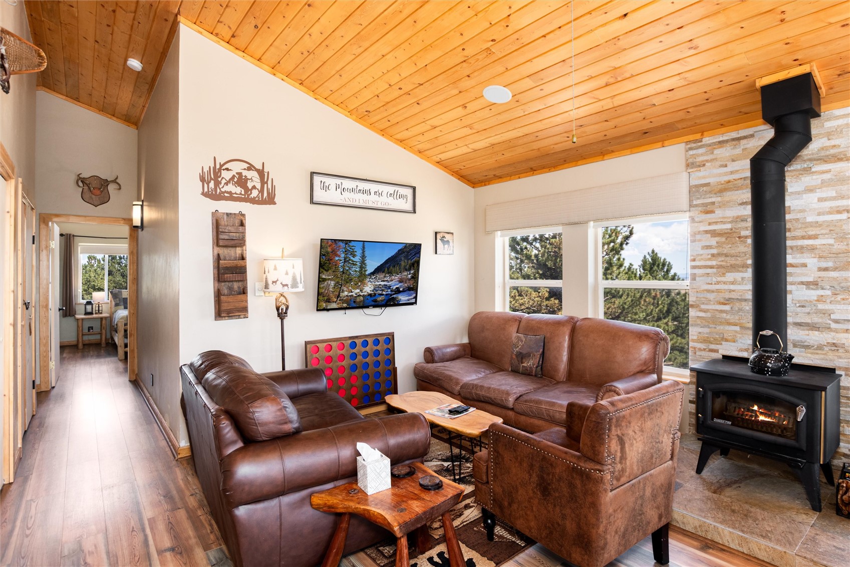 Living area featuring a vaulted wood-finish ceiling, a stone-clad wall with a black wood-burning stove, large windows, and wood-finish flooring