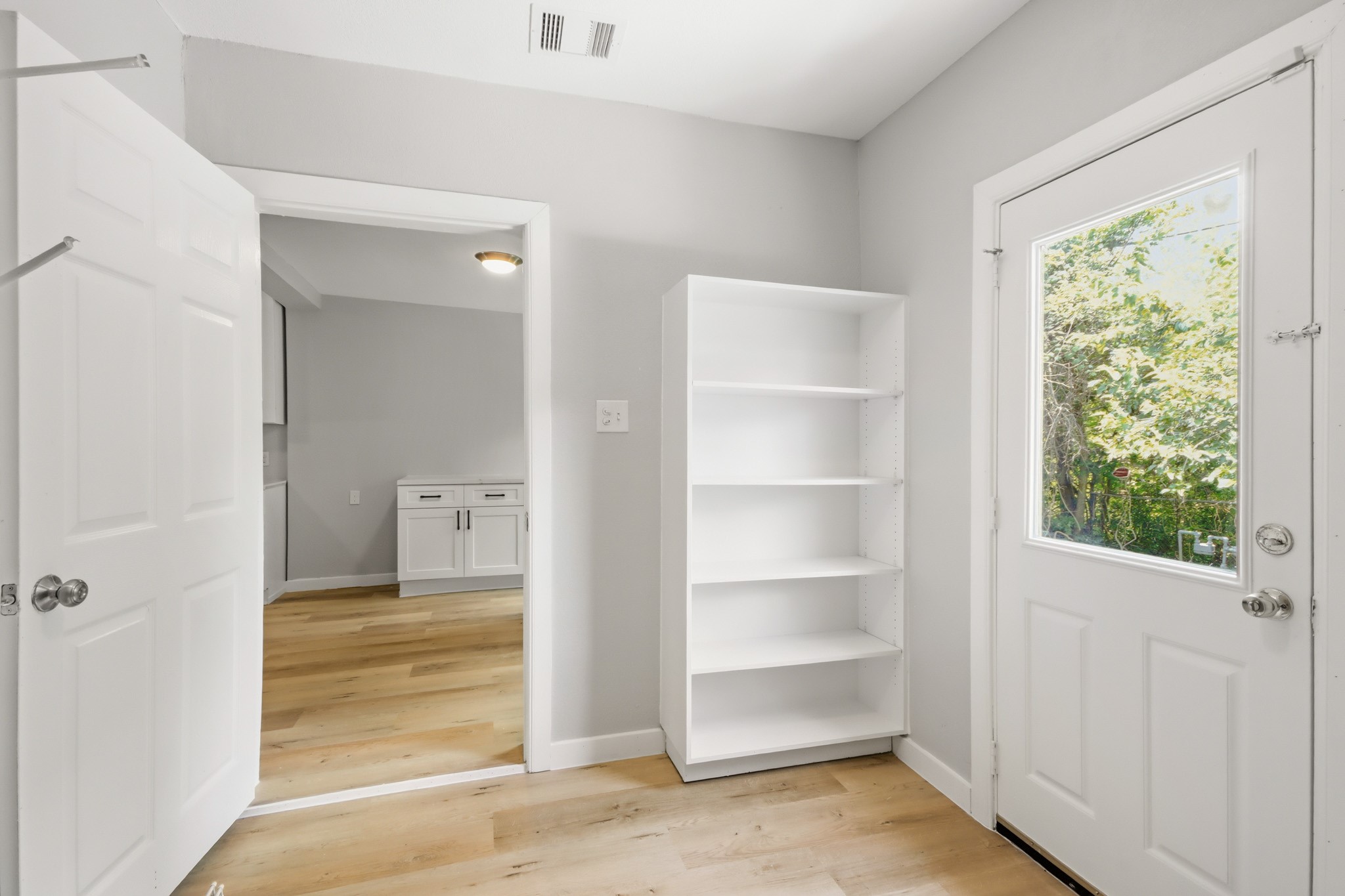 10430 Wiggins Street Houston, TX 77029 - Photo 24 of 27 This room features light wood flooring, neutral gray walls, and a white door with a window leading outside. It includes a white shelving unit and an open doorway to an adjoining space with cabinetry.