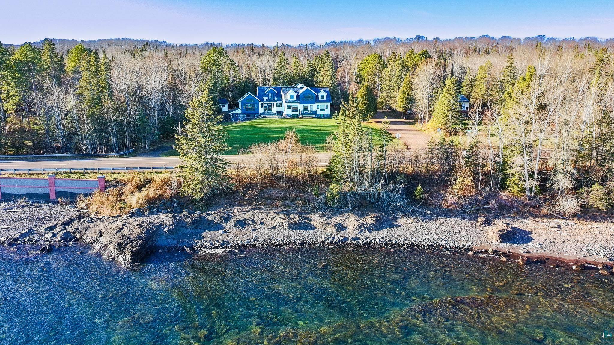 5163 Greenwood Road Duluth, MN 55804 - Photo 2 of 10 Waterfront property featuring a prominent blue and white residence, a verdant lawn, and a dense wooded perimeter