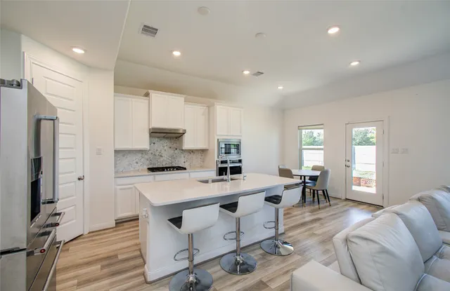 a large white dining hall with stainless steel appliances kitchen island granite countertop a refrigerator a stove a dining table and chairs with wooden floor