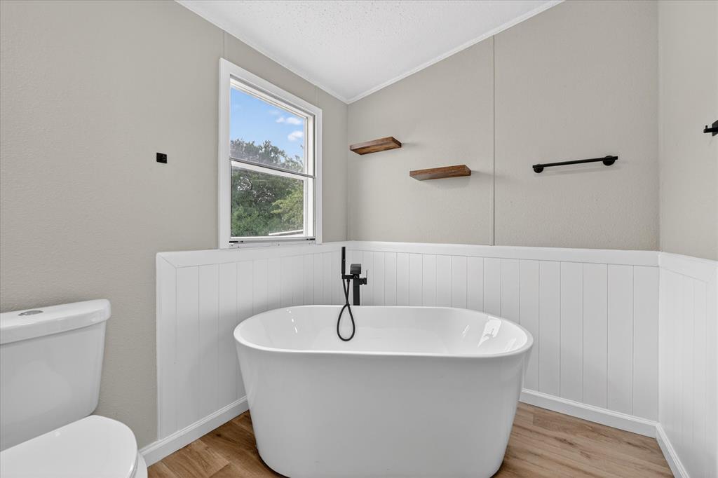 134 Classic Country Court Springtown, TX 76082 - Photo 27 of 35 Bathroom featuring a freestanding soaking tub with a matte black floor-mounted faucet, white wainscoting, wood-finish flooring, and a window providing natural light