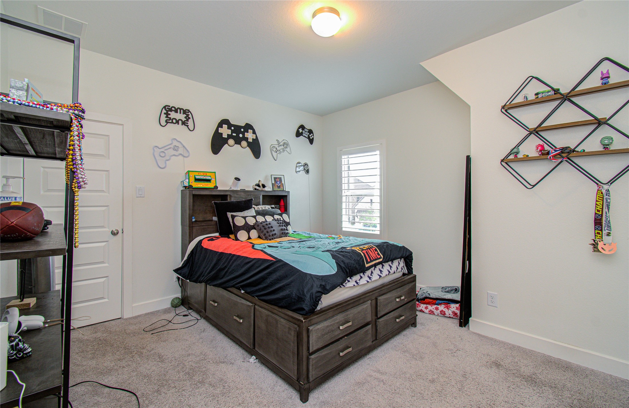 4211 Limestone Terrace Manvel, TX 77578 - Photo 27 of 33 Cozy bedroom with carpeted flooring, featuring a bed with storage drawers underneath, playful gaming-themed wall decorations, and modern shelving. Bright and well-lit with a window for natural light. Ideal for a child's room or guest space.