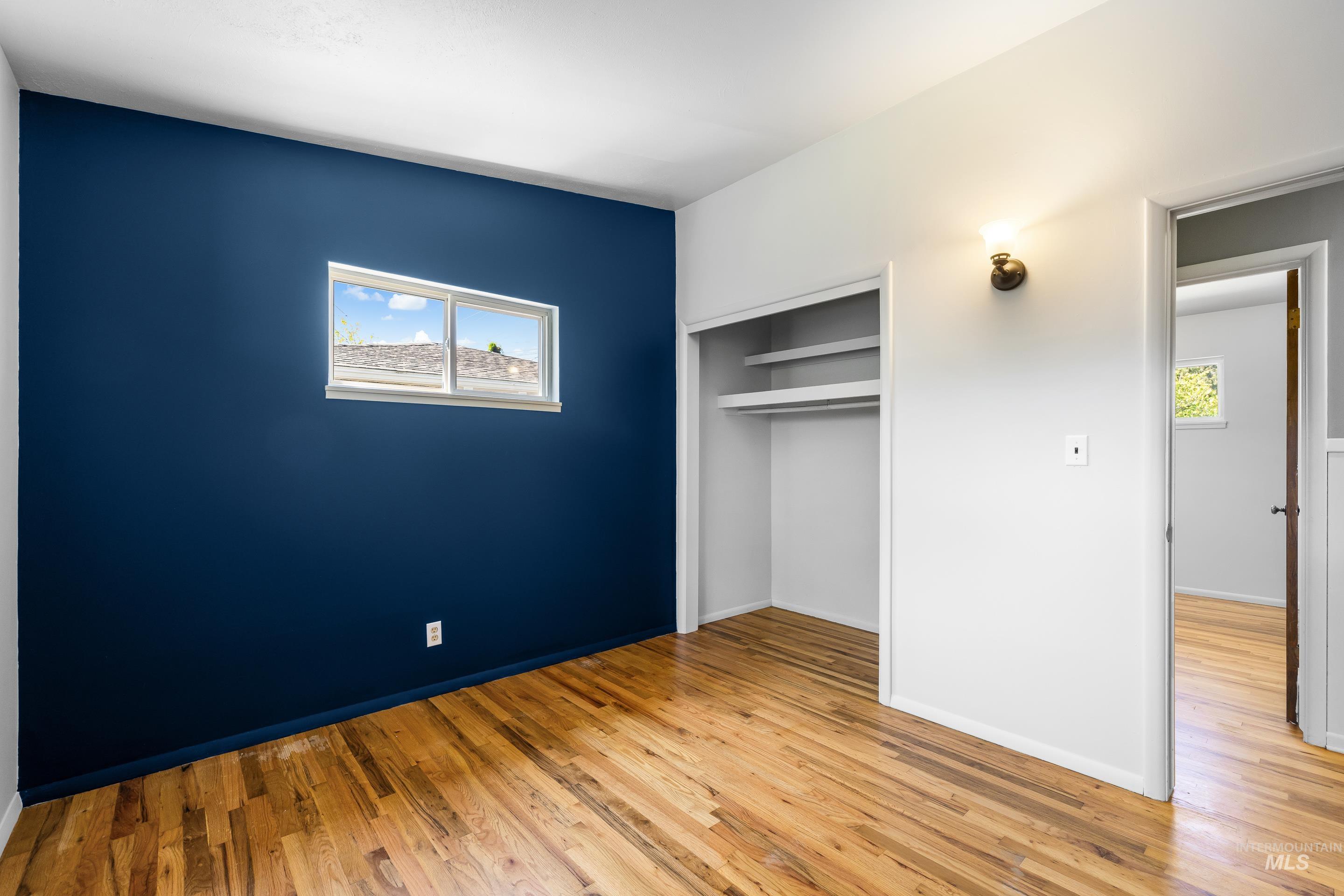 708 South Phillippi Street Boise, ID 83705 - Photo 14 of 25 Room featuring wood-finish flooring, a vibrant blue accent wall, a rectangular window with exterior views, an open closet with shelving, and a wall-mounted light fixture