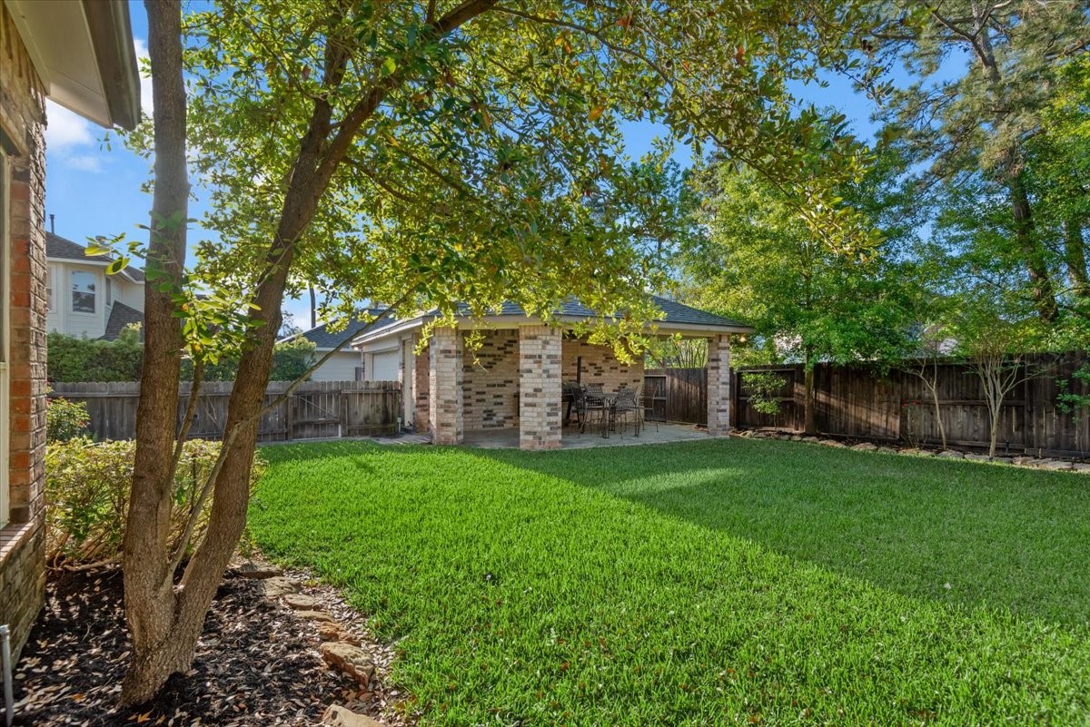 6507 Quiet Pointe Drive Spring, TX 77389 - Photo 29 of 45 This photo showcases a serene backyard with lush green grass, a mature tree, and a charming brick patio area perfect for outdoor dining and relaxation. The space is enclosed by a wooden fence, offering privacy and a cozy atmosphere.