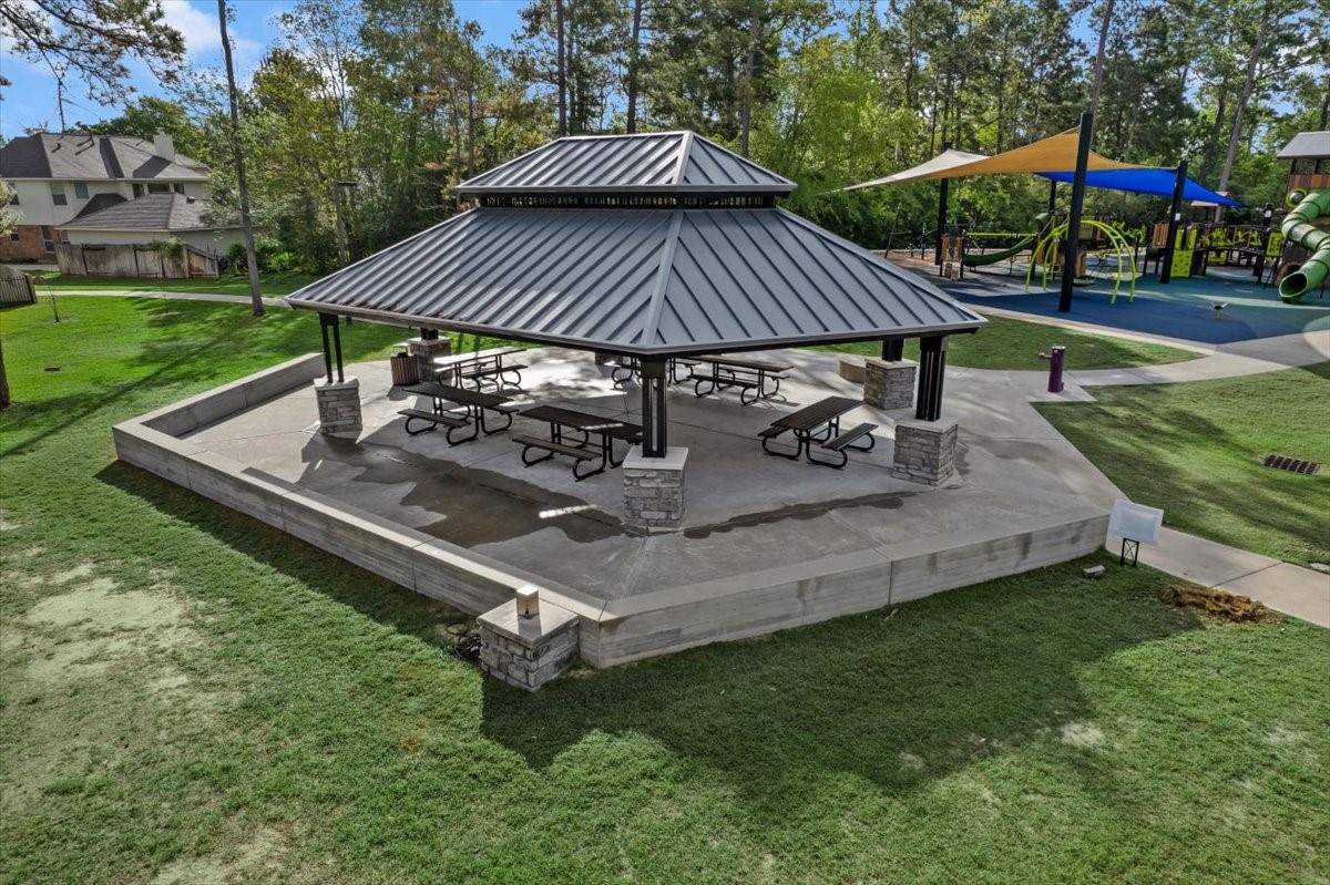 6507 Quiet Pointe Drive Spring, TX 77389 - Photo 33 of 45 This photo features a spacious outdoor pavilion with a metal roof and picnic tables, set on a concrete platform. It's surrounded by a well-maintained lawn, with a nearby playground, offering a perfect spot for gatherings and family activities in a community setting.
