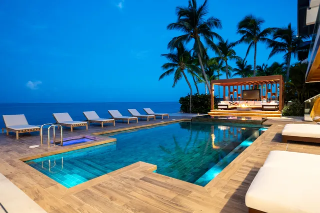 a view of a white building with swimming pool lawn chairs potted plants and palm trees