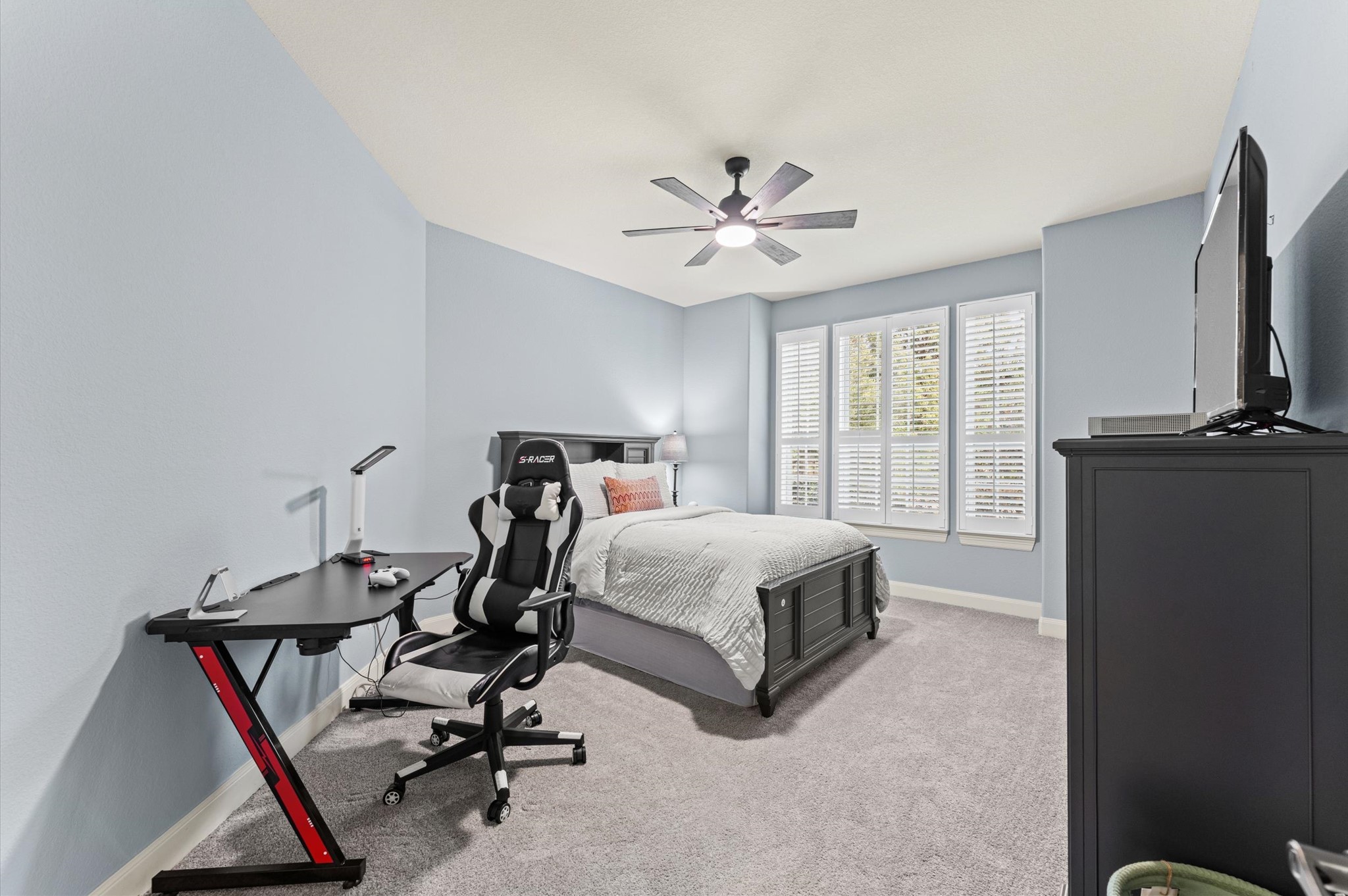 70 Valera Ridge Drive Spring, TX 77389 - Photo 27 of 37 This bedroom features soft blue walls, a cozy carpet, and ample natural light from large windows with shutters. It includes a modern ceiling fan, a comfortable bed, a sleek gaming desk, and a TV on a dresser, creating a functional and inviting space.