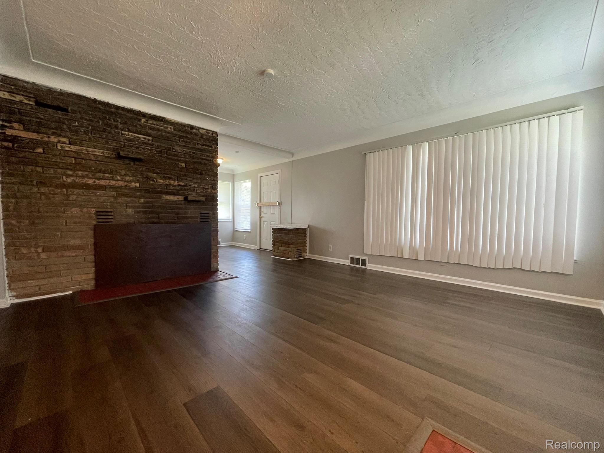 19800 Berg Road Detroit, MI 48219 - Photo 6 of 13 Spacious room featuring wood-finish flooring, a brick accent wall with a firebox, large window with vertical blinds, and neutral wall paint