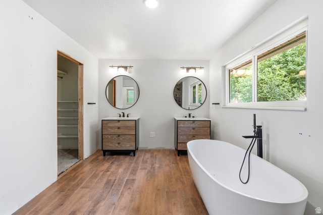 1410 Sumac Drive Logan, UT 84321 - Photo 15 of 28 Bathroom featuring a freestanding white soaking tub with a matte black floor-mounted faucet, wood-finish flooring, dual vanity stations with integrated sinks and round mirrors, and a wide window
