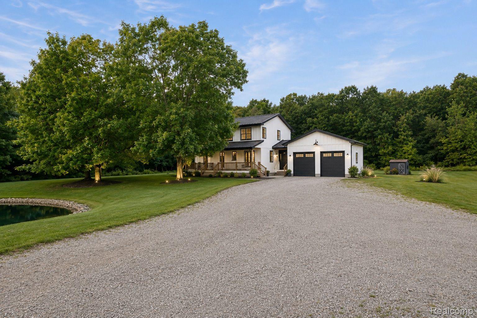 76565 McFadden Road Armada, MI 48005 - Photo 80 of 95 Modern farmhouse style exterior, two-car garage with black doors, gravel driveway, pond feature, and mature trees
