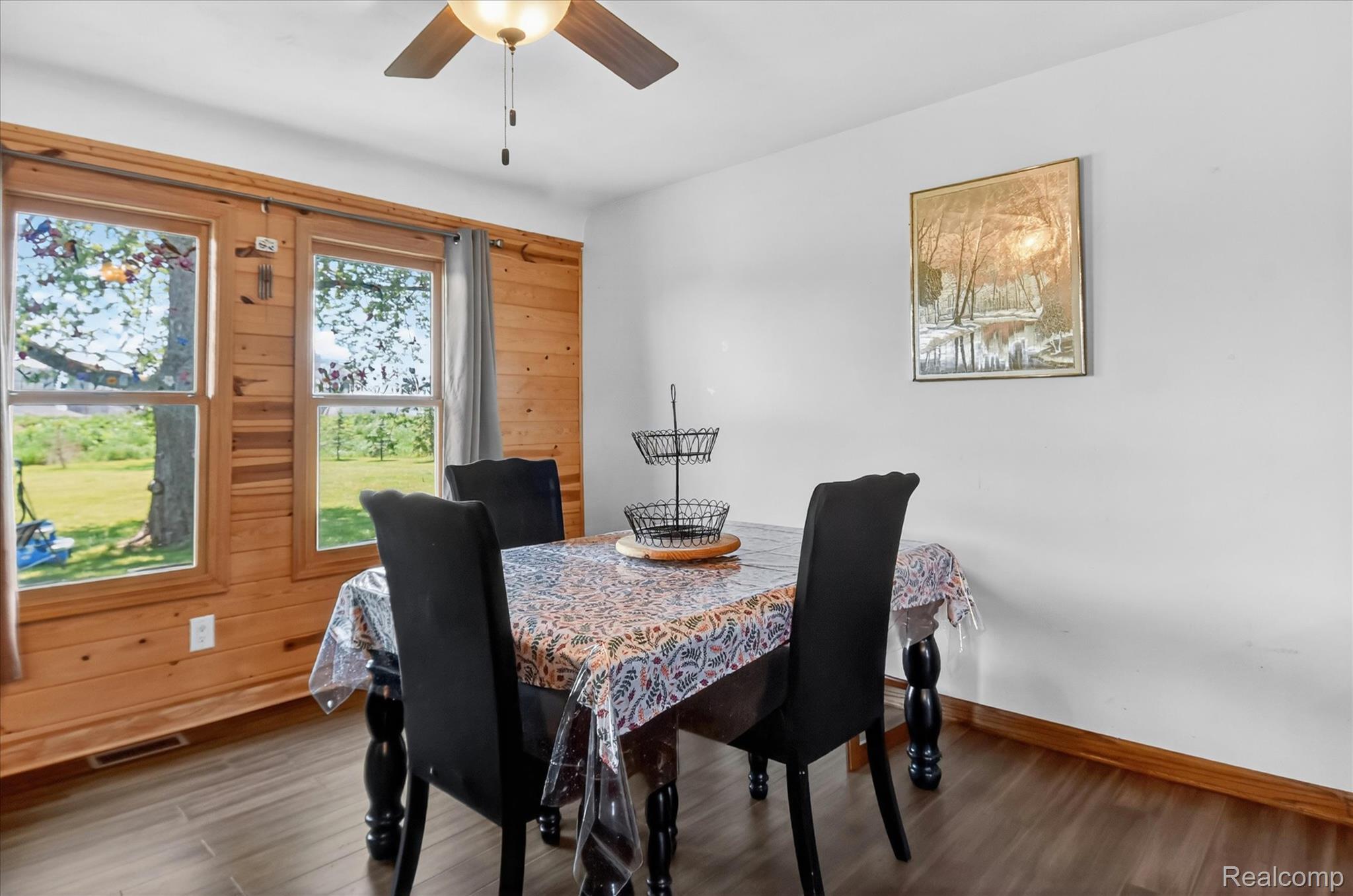 3949 West Burns Line Road Brown City, MI 48416 - Photo 9 of 70 Dining area featuring a wood-panel accent wall, two large windows, wood-finish flooring, and a ceiling fan with integrated lighting