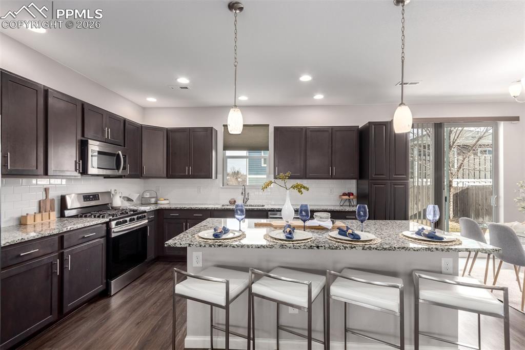 7956 Osage Street Denver, CO 80221 - Photo 11 of 48 Kitchen featuring a breakfast bar, dark brown cabinetry, appliances with stainless steel finishes, hanging light fixtures, and light stone countertops