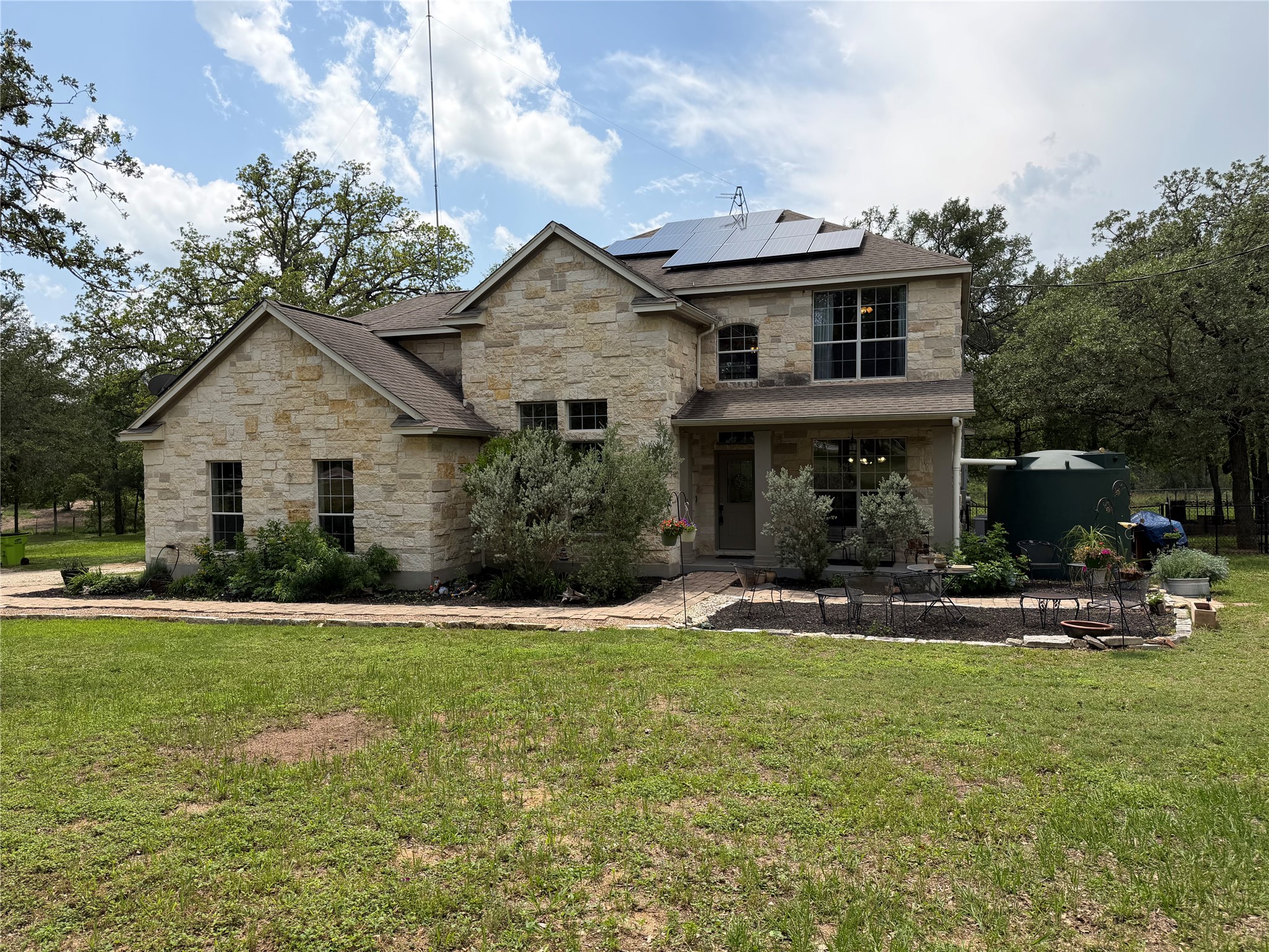 202 The Oaks Boulevard Elgin, TX 78621 - Photo 2 of 40 Two-story stone exterior home featuring multiple solar panels on the roof, a patio area, and a large rainwater collection tank