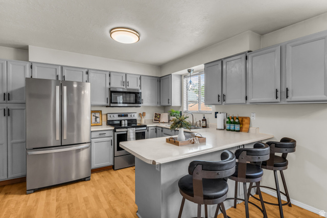 8029 Old Dairy Road Sandy, UT 84094 - Photo 8 of 37 Kitchen featuring light gray cabinetry, stainless steel appliances, light-toned countertops, wood-finish flooring, and a built-in breakfast bar