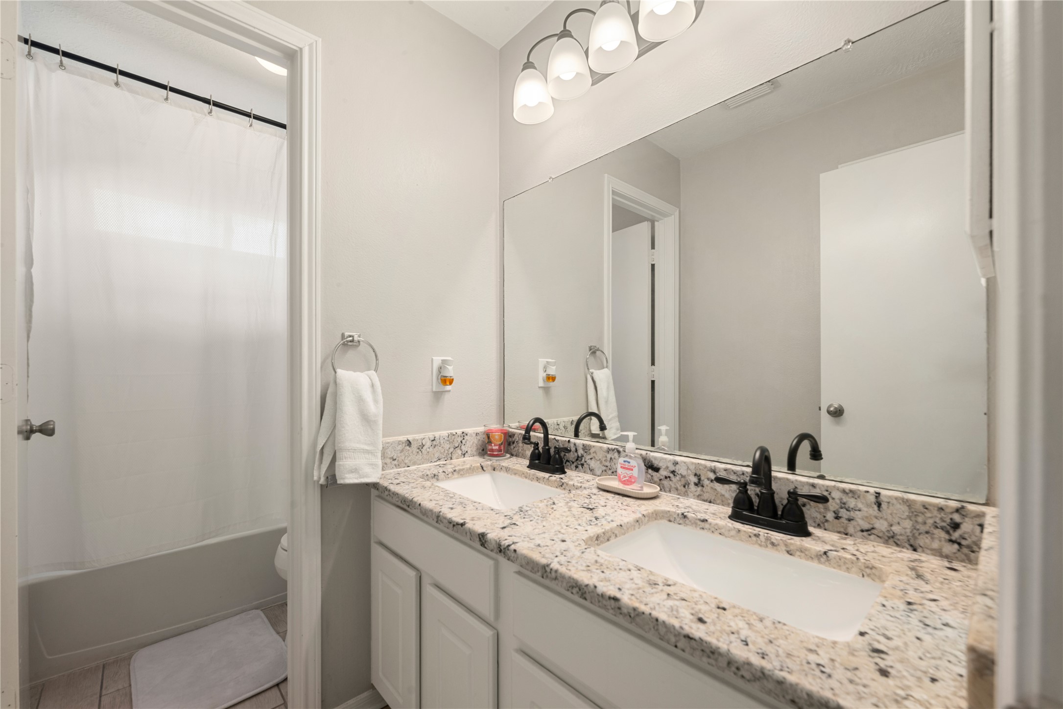 19726 Teller Boulevard Spring, TX 77388 - Photo 19 of 24 This bright and modern secondary bathroom is designed for both style and efficiency. It features a spacious double vanity with granite-style countertops, white cabinetry, and sleek dark hardware. A large mirror and multi-bulb lighting fixture keep the space feeling open and airy, while the separate tub and shower area provides a functional layout for guests or a busy household.