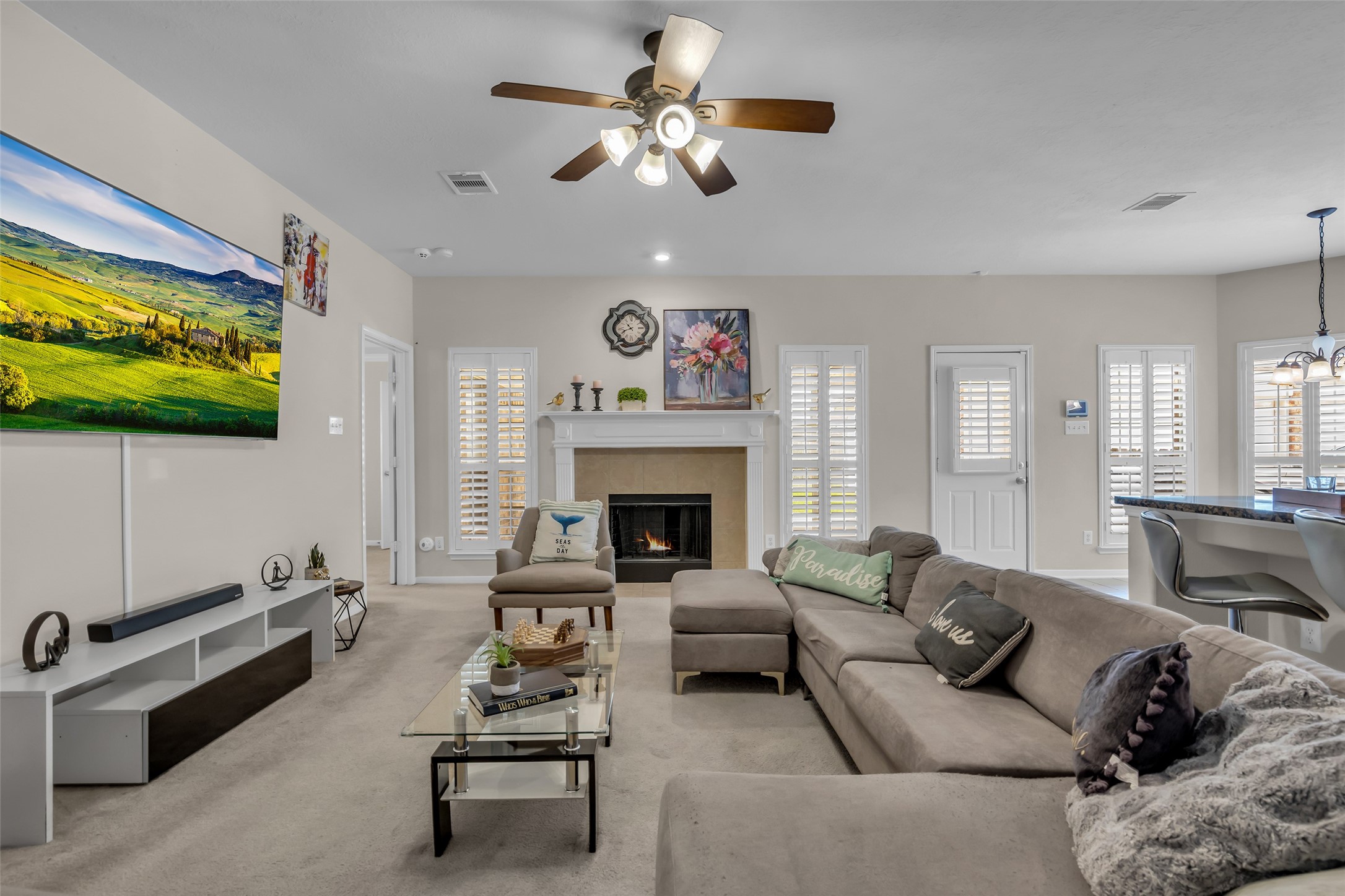 6807 Chapelfield Lane Houston, TX 77049 - Photo 21 of 42 This inviting living room features a large sectional sofa, a cozy fireplace, and a mounted TV. The open layout includes a dining area with a modern light fixture, and ample natural light streams in through multiple windows with shutters. Neutral tones and tasteful decor create a warm, comfortable atmosphere.