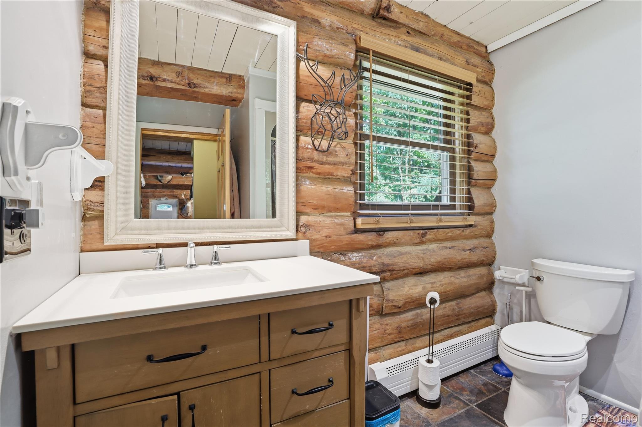 14830 Sheridan Road Byron, MI 48418 - Photo 24 of 74 Bathroom featuring a log wall, white vanity with integrated sink, white countertop, and a framed mirror