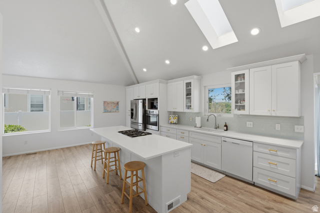 5788 Walden Ridge Drive Murray, UT 84123 - Photo 2 of 29 Kitchen featuring a vaulted ceiling with skylights, wood-finish flooring, white cabinetry, a large center island with a built-in cooktop, and stainless steel appliances
