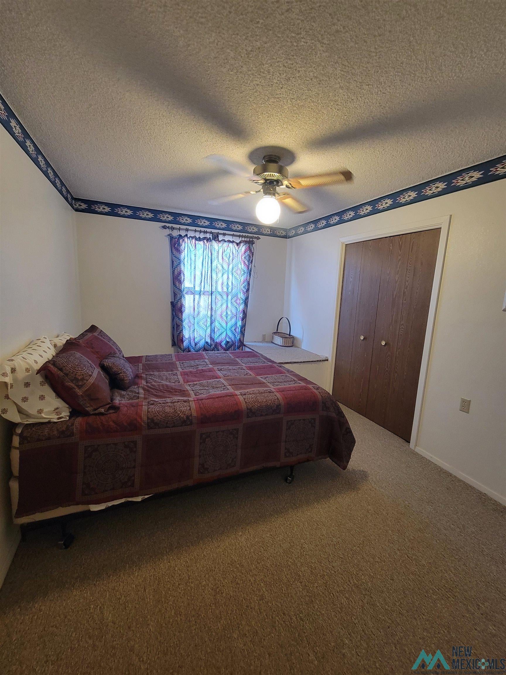 501 Springland Boulevard Elephant Butte, NM 87935 - Photo 12 of 38 Interior room featuring textured carpeting, a window with patterned curtains, a ceiling fan with light fixture, and wood-finish bi-fold closet doors