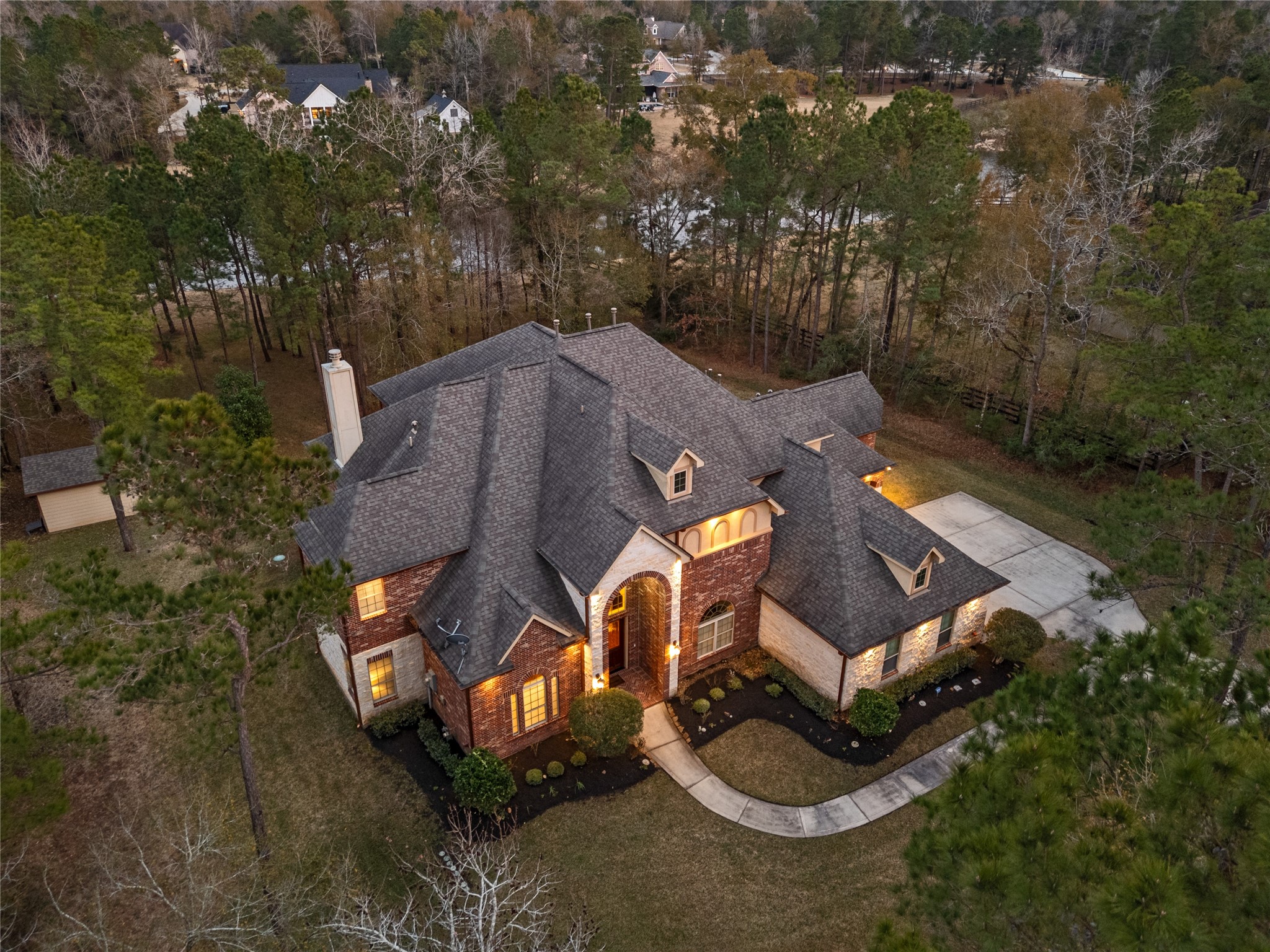 23421 Cannondale Loop Montgomery, TX 77316 - Photo 40 of 47 Stunning aerial view at dusk highlighting the home’s elegant brick and stone façade, arched entryway, and welcoming exterior lighting. Nestled among mature trees, the property offers a peaceful, private setting while showcasing a circular-style driveway approach and expansive roofline that emphasize the home’s impressive scale and presence.