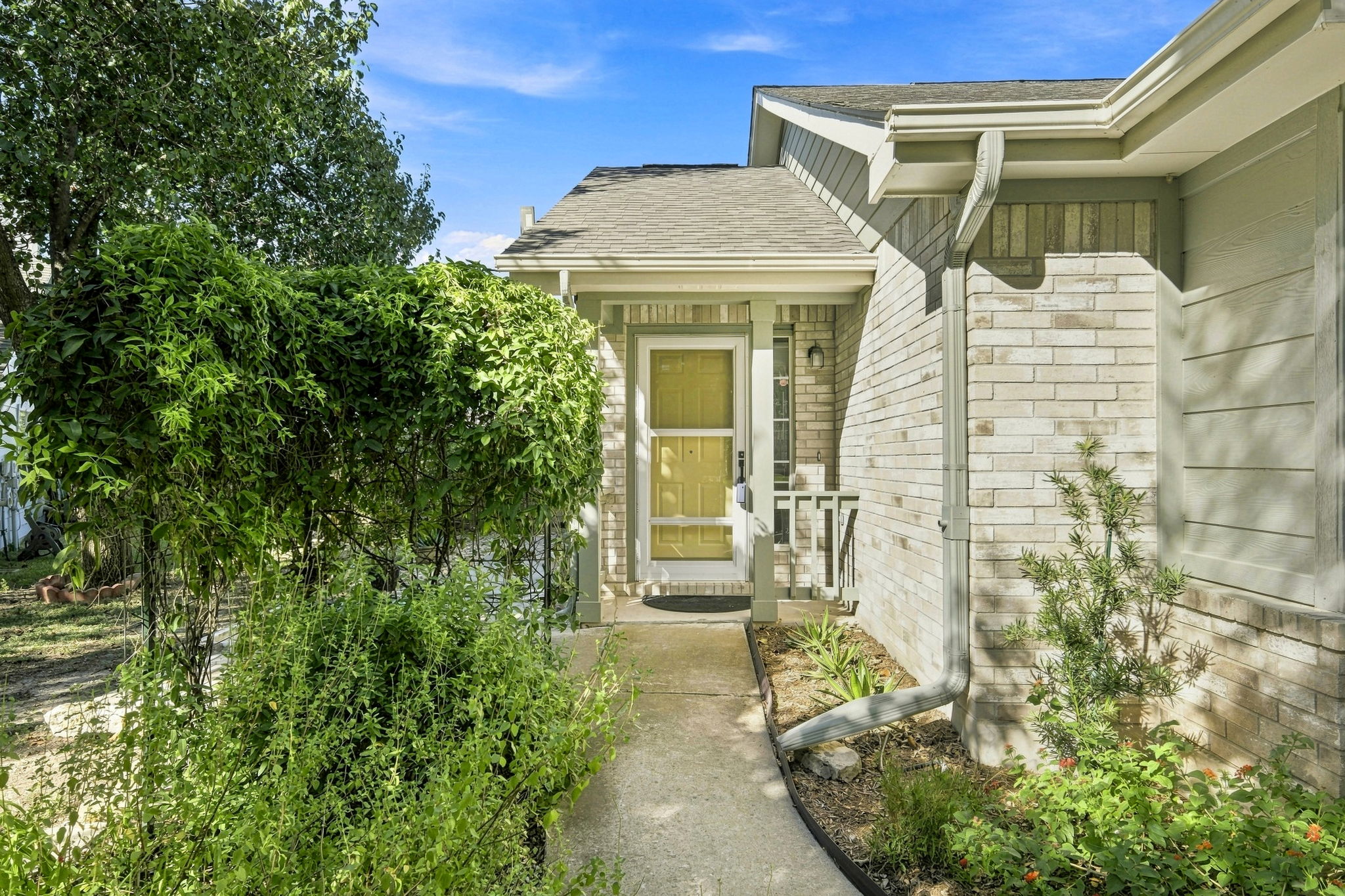 A concrete walkway leads past lush greenery to the covered front entrance of this home.