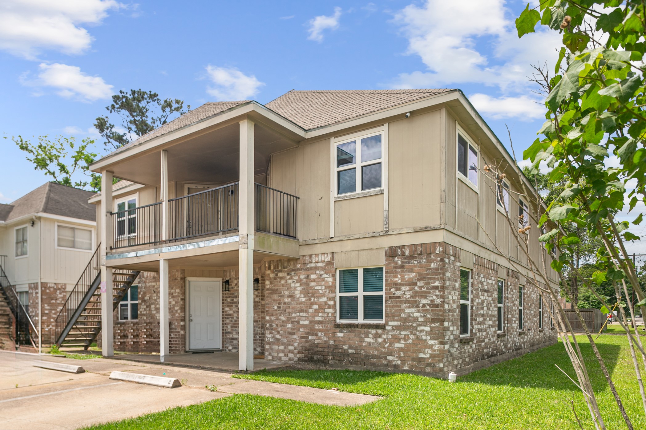 This is a two-story home with a mix of brick and siding exterior. It features a covered balcony on the upper level and an accessible entrance below. The property has a small grassy area and is situated in a residential area with a clear sky backdrop.