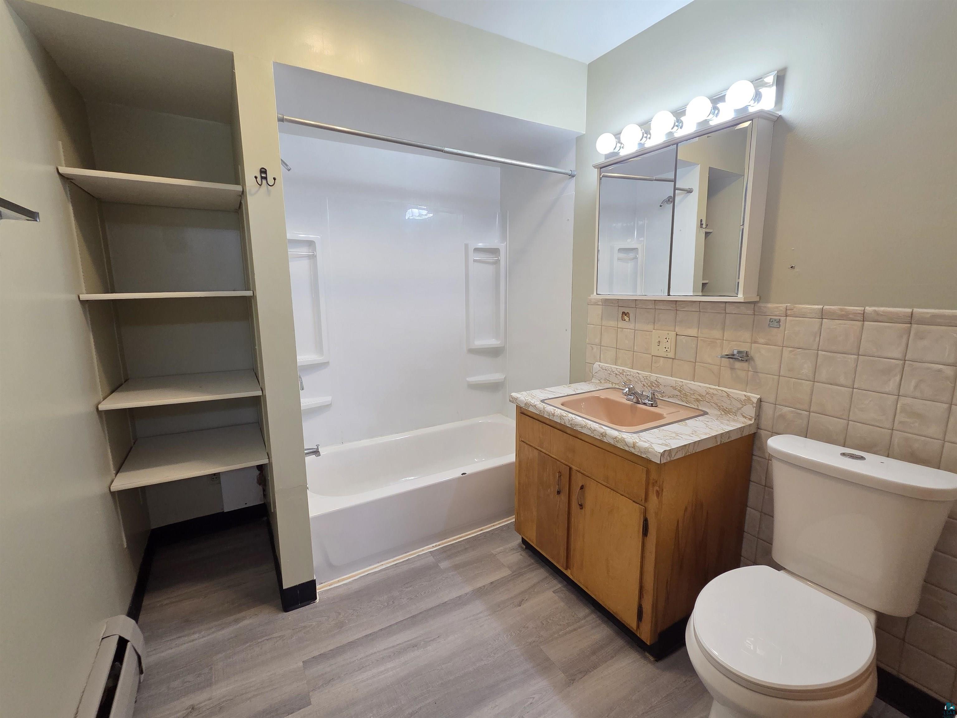 56 East 2nd Street Superior, WI 54880 - Photo 10 of 18 Bathroom featuring wood-finish flooring, built-in shelving, a full-size bathtub with shower, a vanity with a counter and integrated sink, and a commode