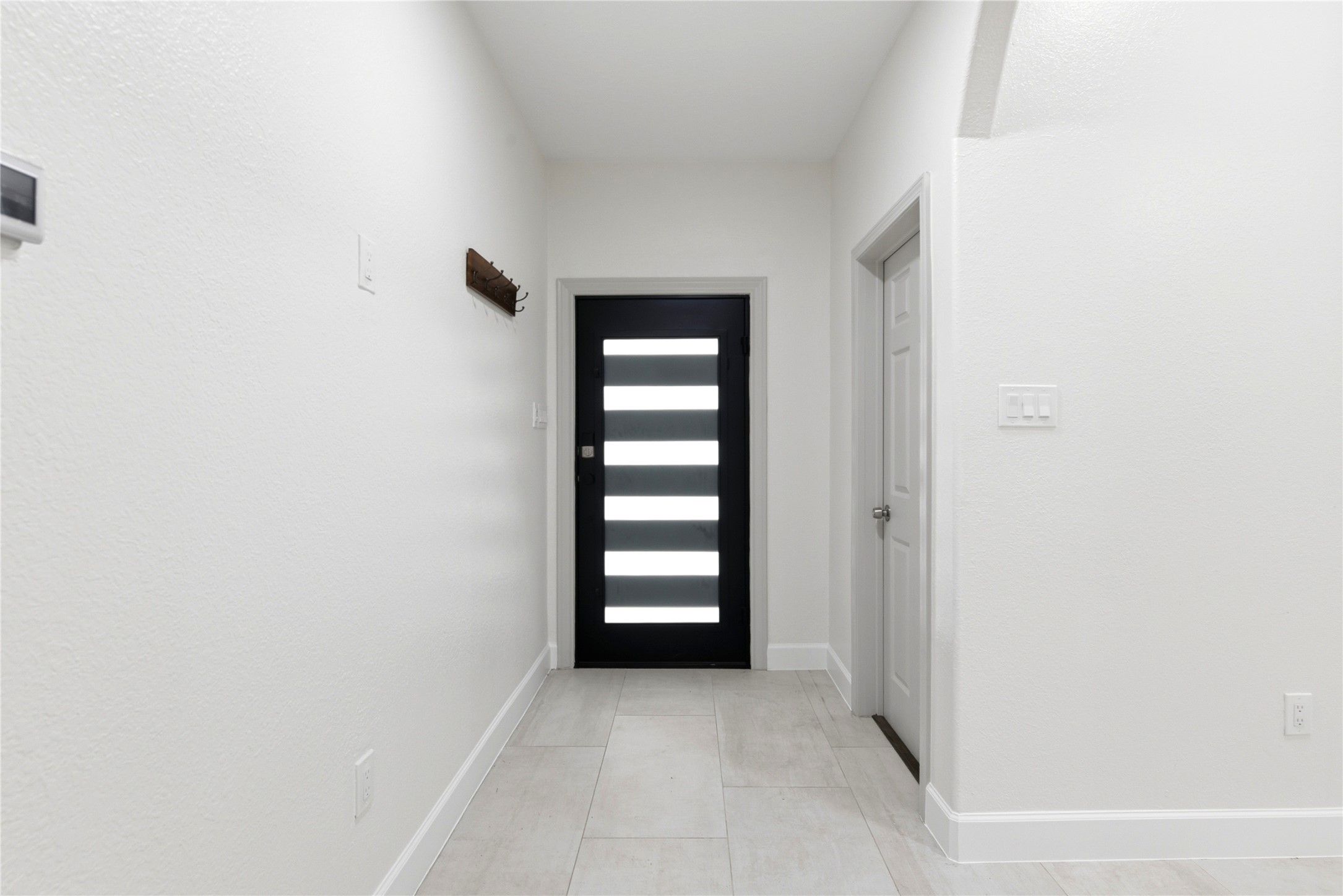 9403 Spaulding Street Houston, TX 77016 - Photo 7 of 50 This photo shows a modern, minimalist entryway with white walls and light tile flooring. The black front door features horizontal glass panels, allowing natural light inside.