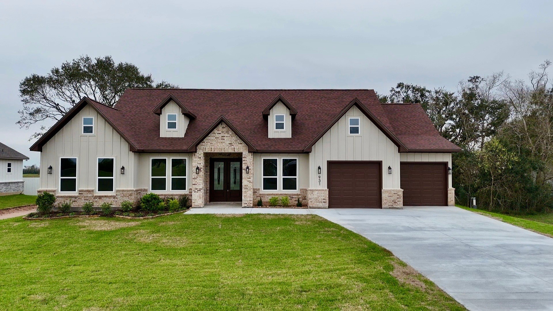 This photo shows a charming house with a rustic design, featuring a red gabled roof and a mix of stone and siding exterior. It has a spacious front yard with a paved driveway leading to an attached two-car garage. Large windows offer plenty of natural light.