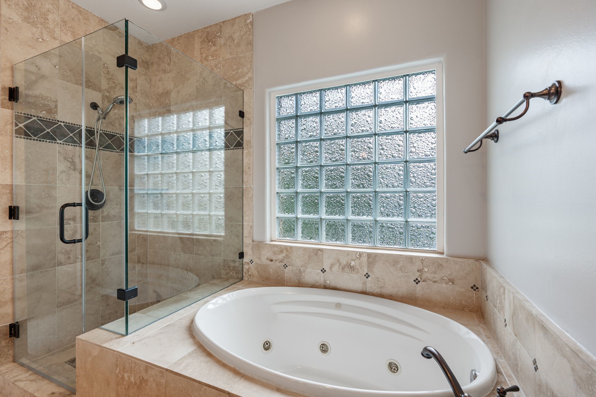 3822 Merrick Street Houston, TX 77025 - Photo 20 of 34 A closer look at the wet area of the primary bath highlights the oversized jetted soaking tub set into travertine tile surround with decorative inlay accents, the large glass block window above it, and the frameless glass shower enclosure featuring a full-height travertine tile surround with decorative mosaic banding and a built-in niche.