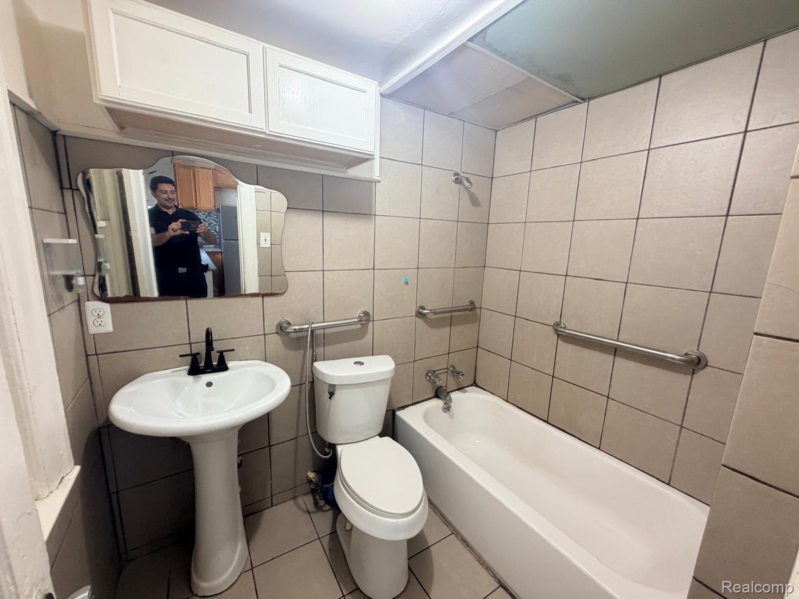 3262 Hanley Street Hamtramck, MI 48212 - Photo 4 of 7 Pedestal sink with a matte black faucet, wall-mounted vanity mirror, white overhead cabinetry, tile flooring, and a shower-tub combination with grab bars