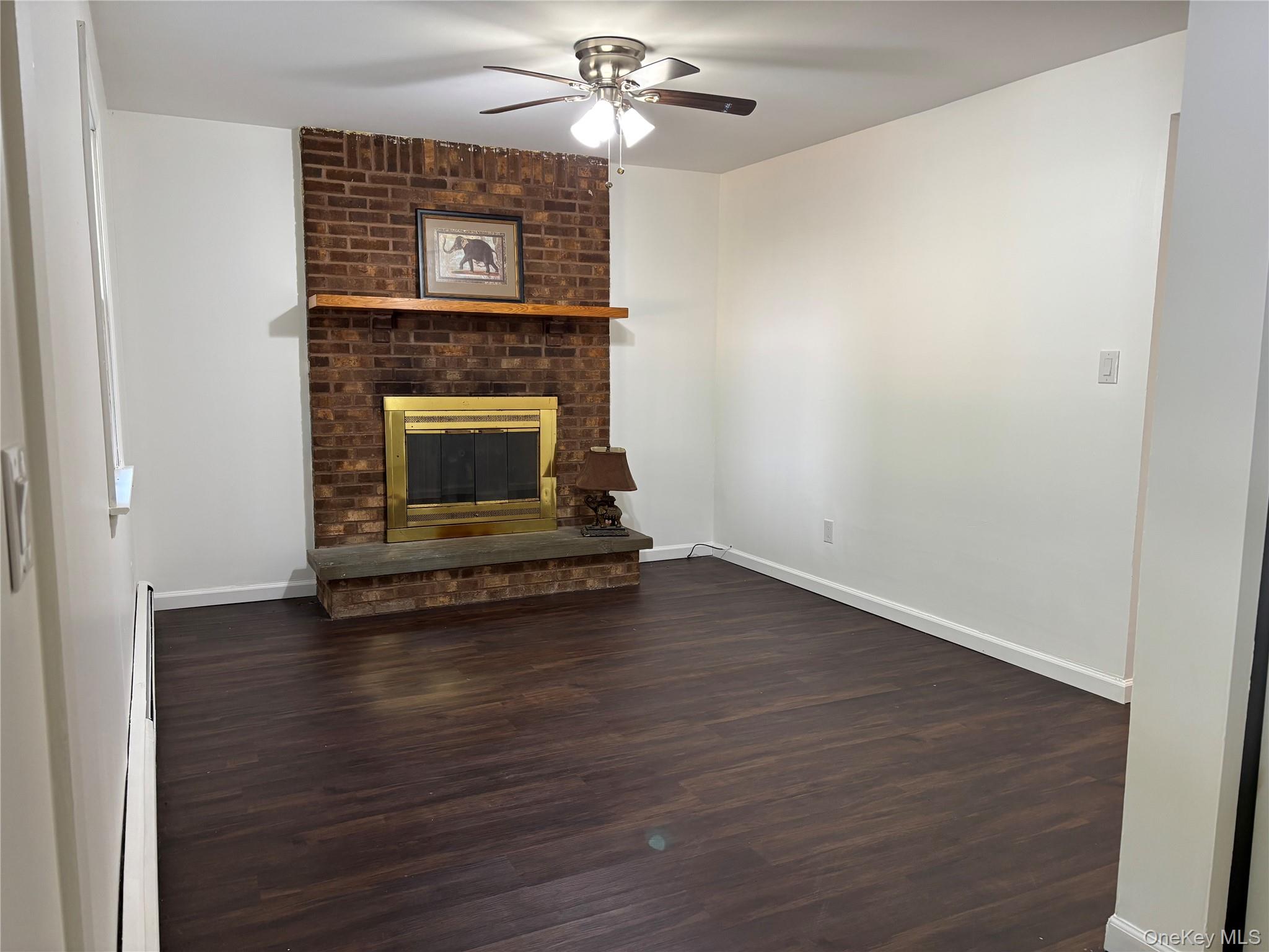 679 A Meadow Road Smithtown, NY 11787 - Photo 5 of 30 Unfurnished living room featuring dark wood-style flooring, a ceiling fan, a baseboard heating unit, and a brick fireplace