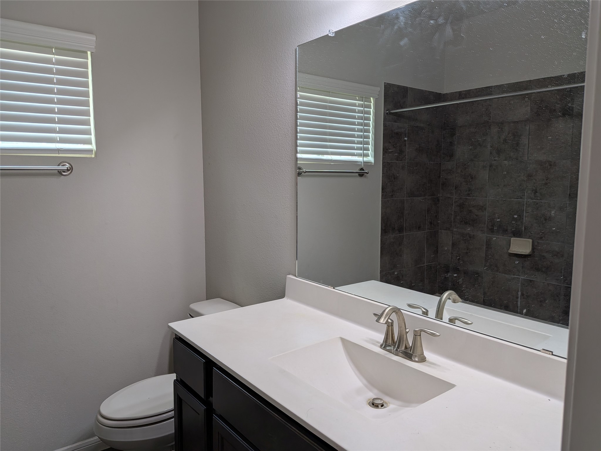 15730 Joe Dimaggio Street Splendora, TX 77372 - Photo 18 of 20 This bathroom features a modern sink with a sleek countertop, a large mirror, and dark cabinetry. It includes a toilet and a shower with dark tile accents, complemented by a window with blinds for natural light.