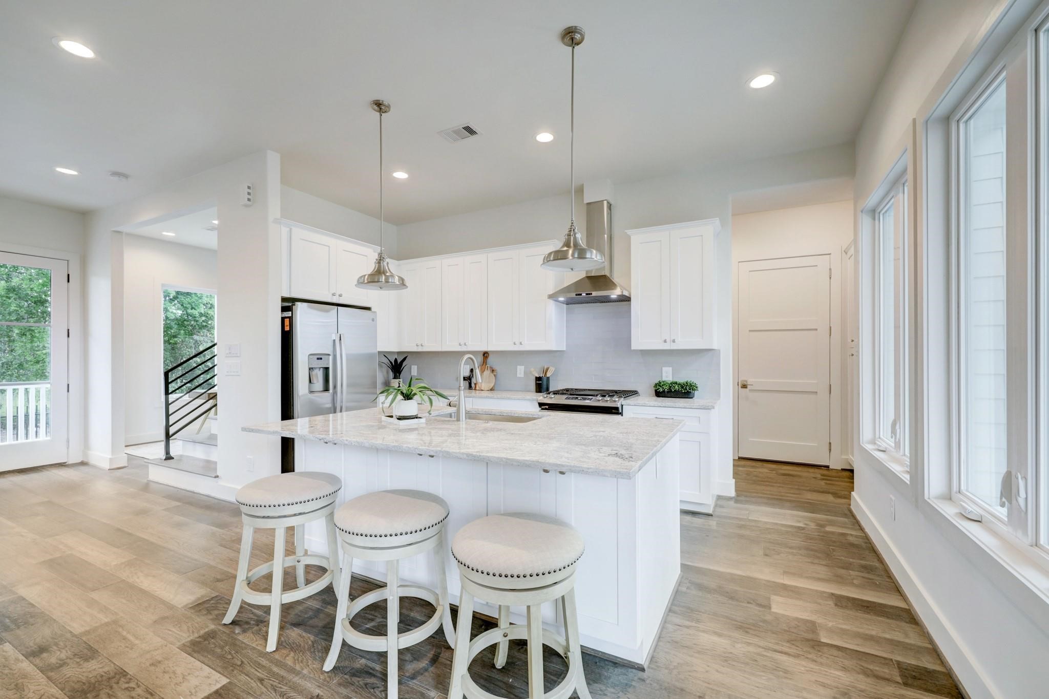 3827 Arlington Street Houston, TX 77018 - Photo 10 of 39 This kitchen features a modern, open design with white cabinetry, a large central island with seating, stainless steel appliances, and ample natural light from multiple windows. The space is complemented by sleek wood flooring and contemporary pendant lighting, creating a bright and inviting atmosphere.