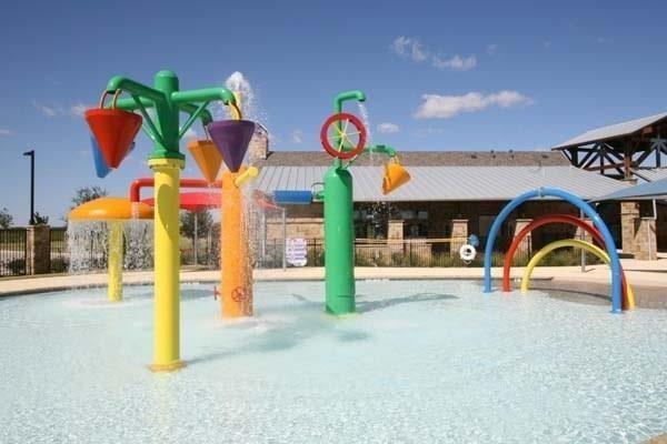 300 Rugby Lane McKinney, TX 75072 - Photo 22 of 25 Outdoor splash pad with multiple colorful water features, including tipping buckets and arch sprays, complemented by a nearby stone-clad building with a metal roof