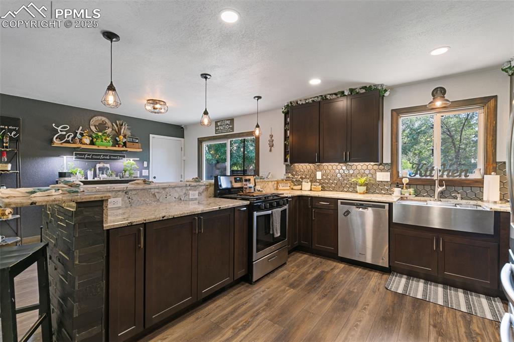 1921 Van Diest Road Colorado Springs, CO 80915 - Photo 7 of 45 Kitchen with dark brown cabinets, a peninsula, appliances with stainless steel finishes, light stone counters, and dark wood-style floors