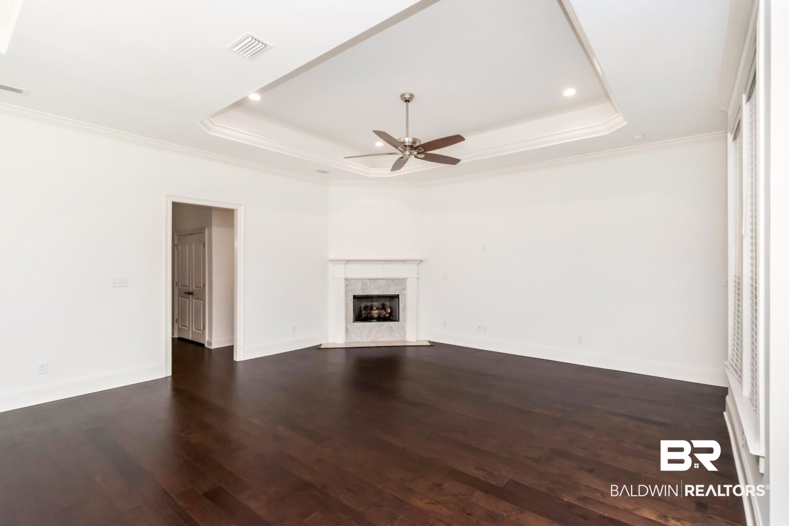 3528 Rue Royal Mobile, AL 36693 - Photo 21 of 53 Spacious room featuring dark wood-finish flooring, a tray ceiling with recessed lighting, a ceiling fan, a fireplace with a stone surround, and white trim throughout