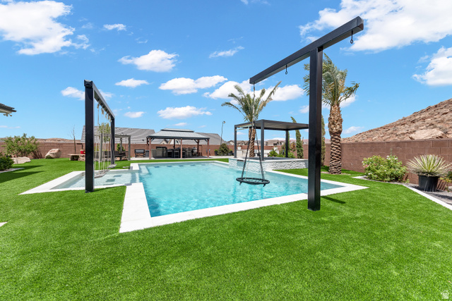 6105 Bunda Way St. George, UT 84790 - Photo 37 of 49 Expansive backyard featuring a rectangular swimming pool with white coping, artificial turf, a built-in pergola structure, mature palm trees, and perimeter fencing
