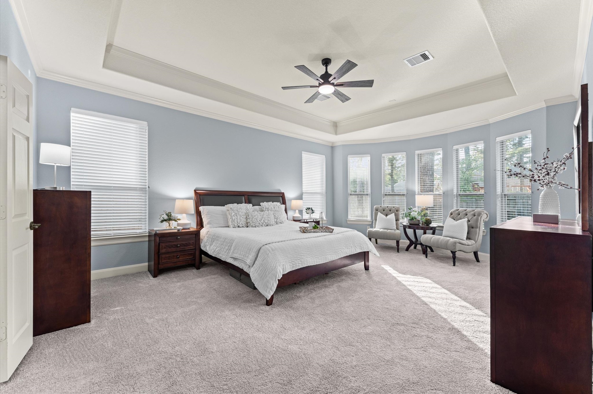 70 Valera Ridge Drive Spring, TX 77389 - Photo 22 of 37 This spacious master bedroom features soft carpeting, a tray ceiling with a ceiling fan, and abundant natural light from multiple large windows. The room is elegantly furnished with a dark wood bed frame, matching nightstands, and a cozy seating area, creating a warm and inviting atmosphere.