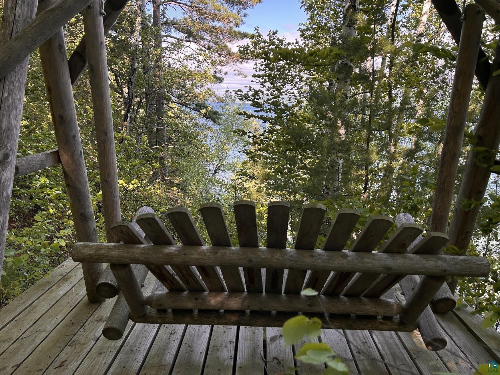 1596 North Shore Road La Pointe, WI 54850 - Photo 21 of 21 Wooden porch swing on a multi-level wood deck, surrounded by mature trees and lush foliage, with a body of water in the distance under a clear sky