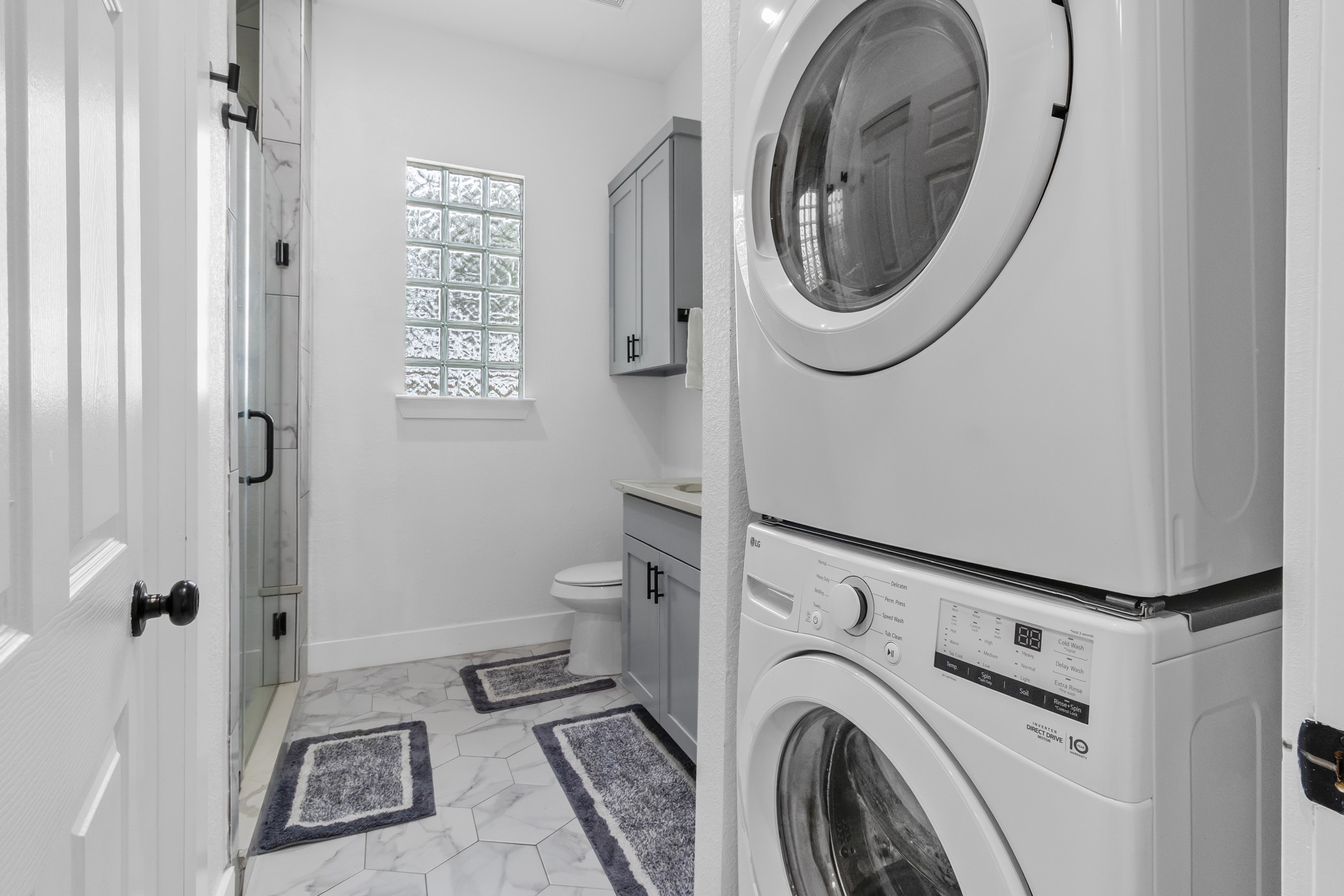 123 Zacate Street Elgin, TX 78621 - Photo 24 of 33 Laundry area with stacked washer and dryer unit, featuring a full bathroom with glass block window, frameless glass shower enclosure with black hardware, hexagon tile flooring, and a vanity with gray cabinetry and integrated sink