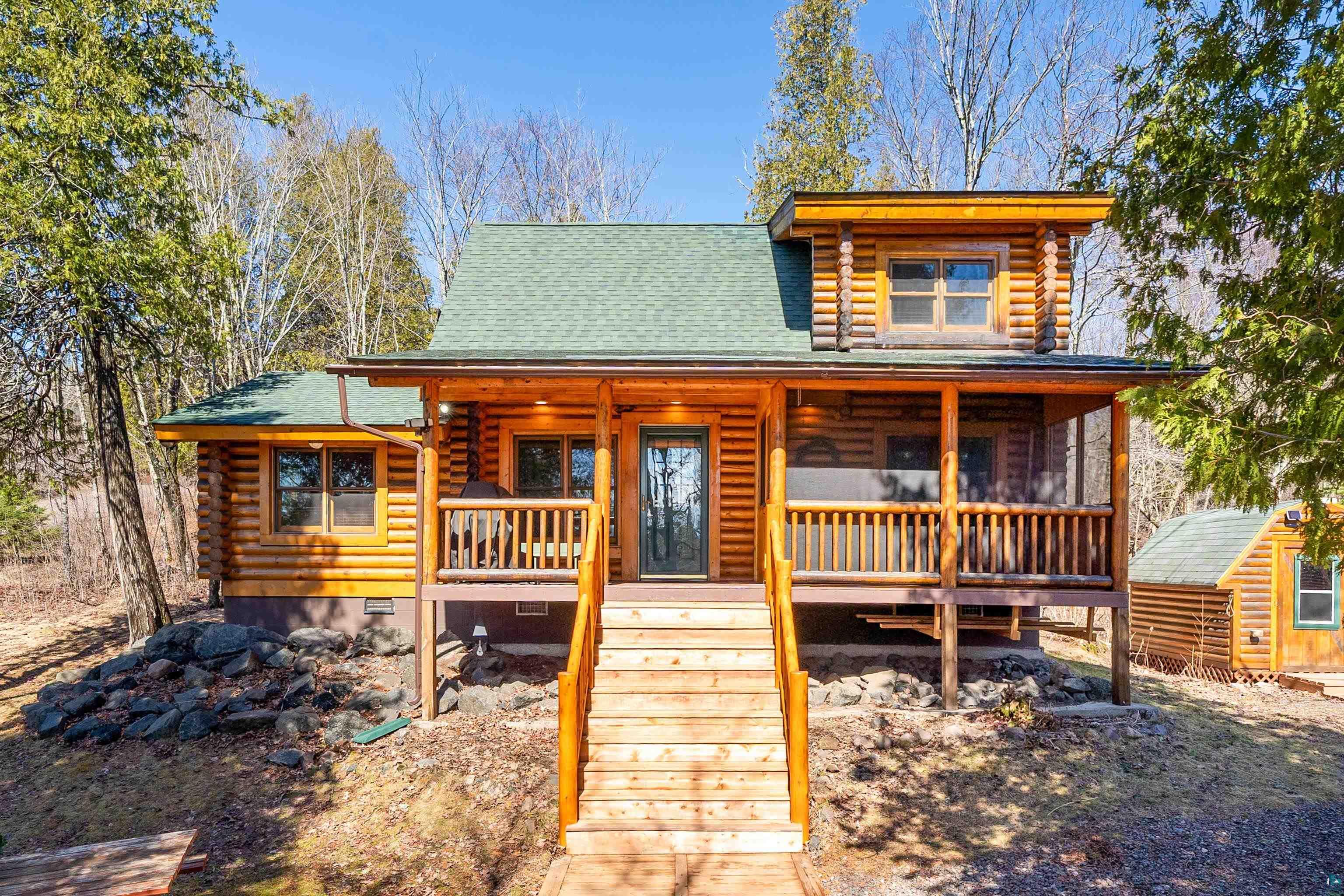 Log cabin exterior featuring a covered porch with natural wood railings, a prominent staircase, and green shingle roofing