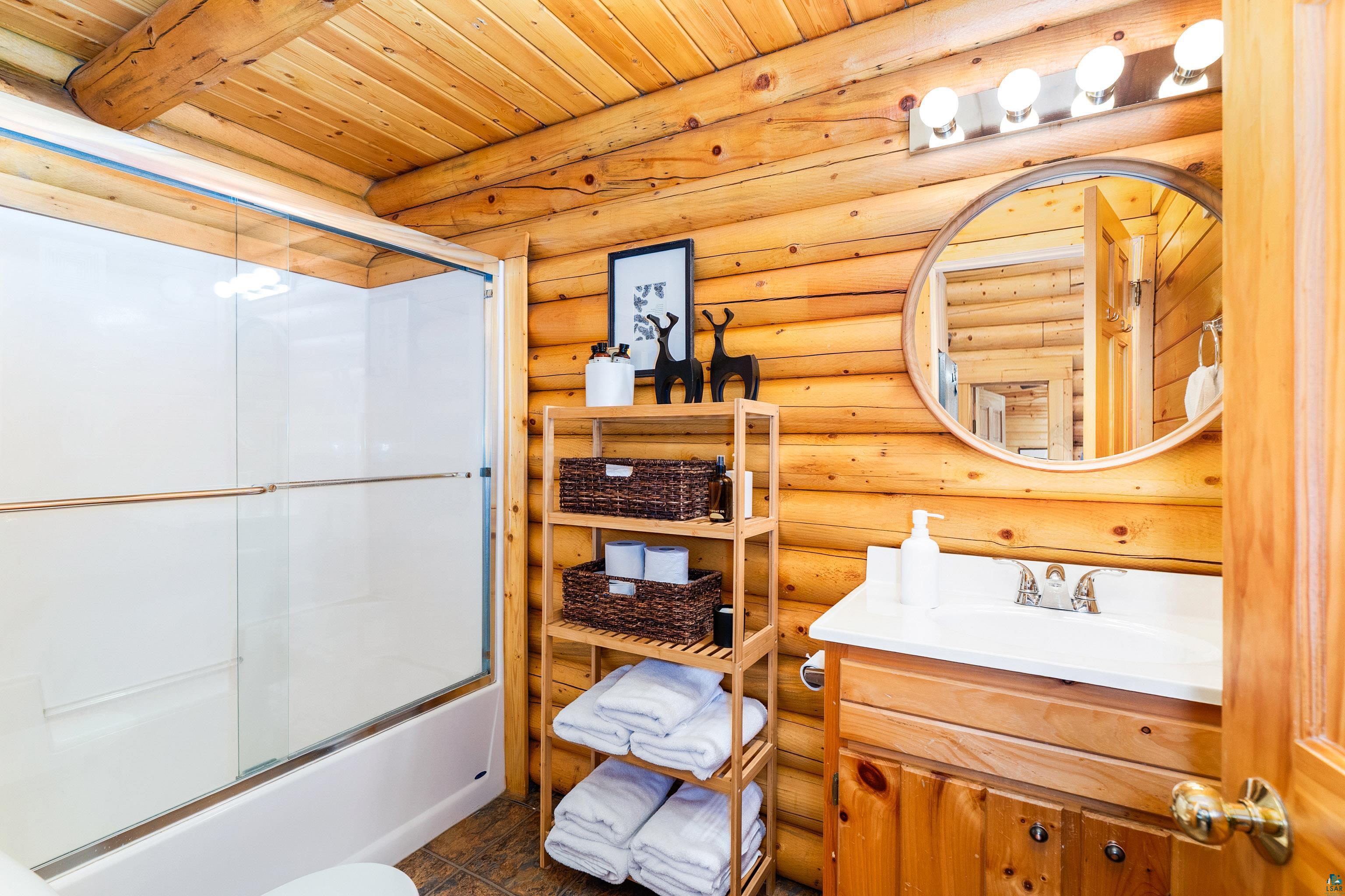 2659 Ridgeview Drive Two Harbors, MN 55616 - Photo 31 of 56 Bathroom featuring log cabin walls and ceiling, a white tub with clear glass sliding shower doors, a wood vanity with a white countertop and integrated sink, and a circular wall mirror with a wood frame