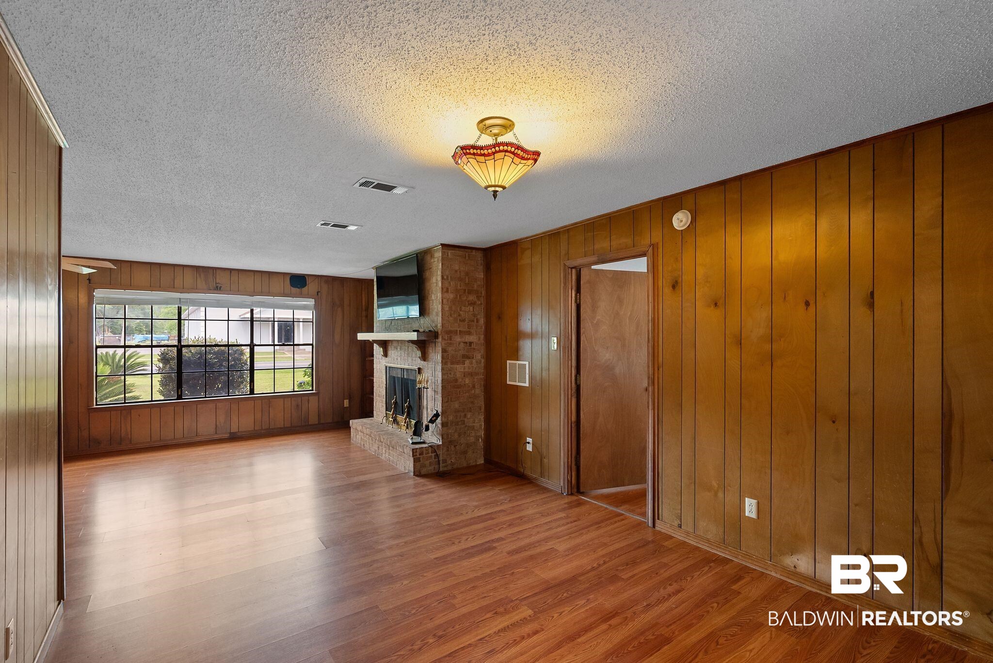 716 North Cedar Street Foley, AL 36535 - Photo 9 of 25 Spacious room featuring wood-finish flooring, wood paneling, a brick fireplace with mantel, and a large window with exterior landscaping visible