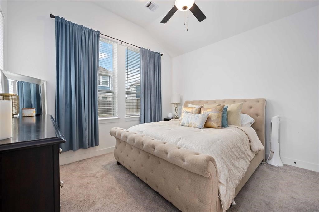 10014 Nectar Path Iowa Colony, TX 77583 - Photo 17 of 26 Cozy bedroom with a plush tufted bed, neutral carpeting, and soft blue curtains. The room features a ceiling fan and ample natural light from double windows, creating a serene and inviting atmosphere.