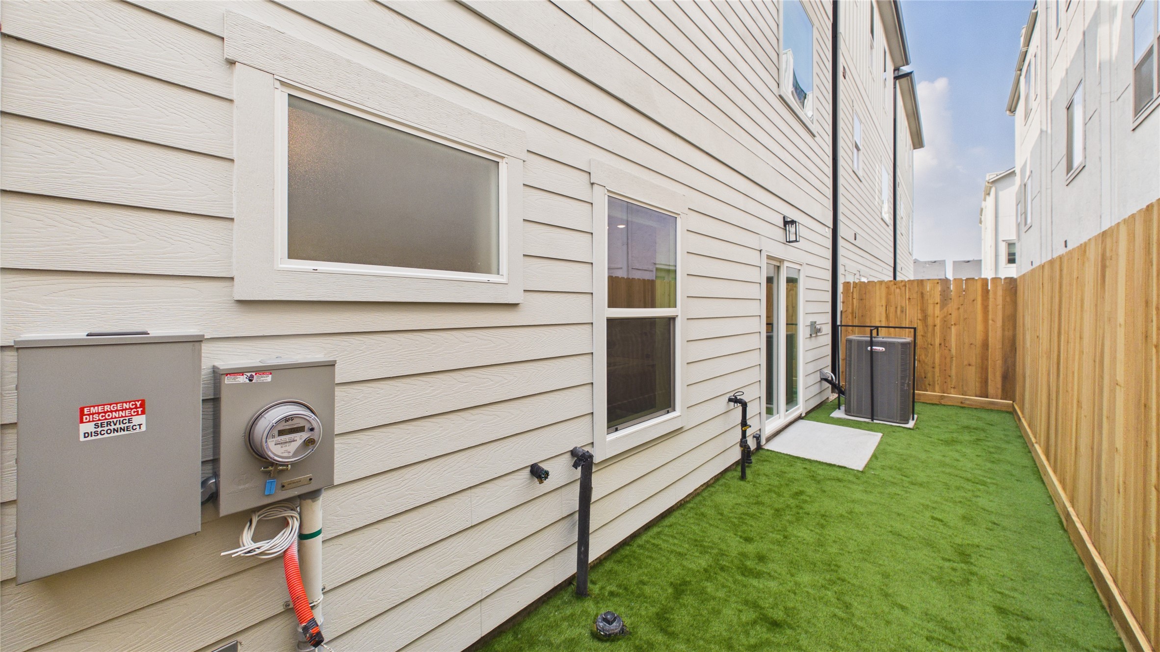 602 Skyland Terrace Way Houston, TX 77009 - Photo 38 of 40 This photo shows a narrow side yard with artificial grass, a wooden fence, and access to the home's utilities. The siding is light-colored, and there are multiple windows and a patio door leading to the area.